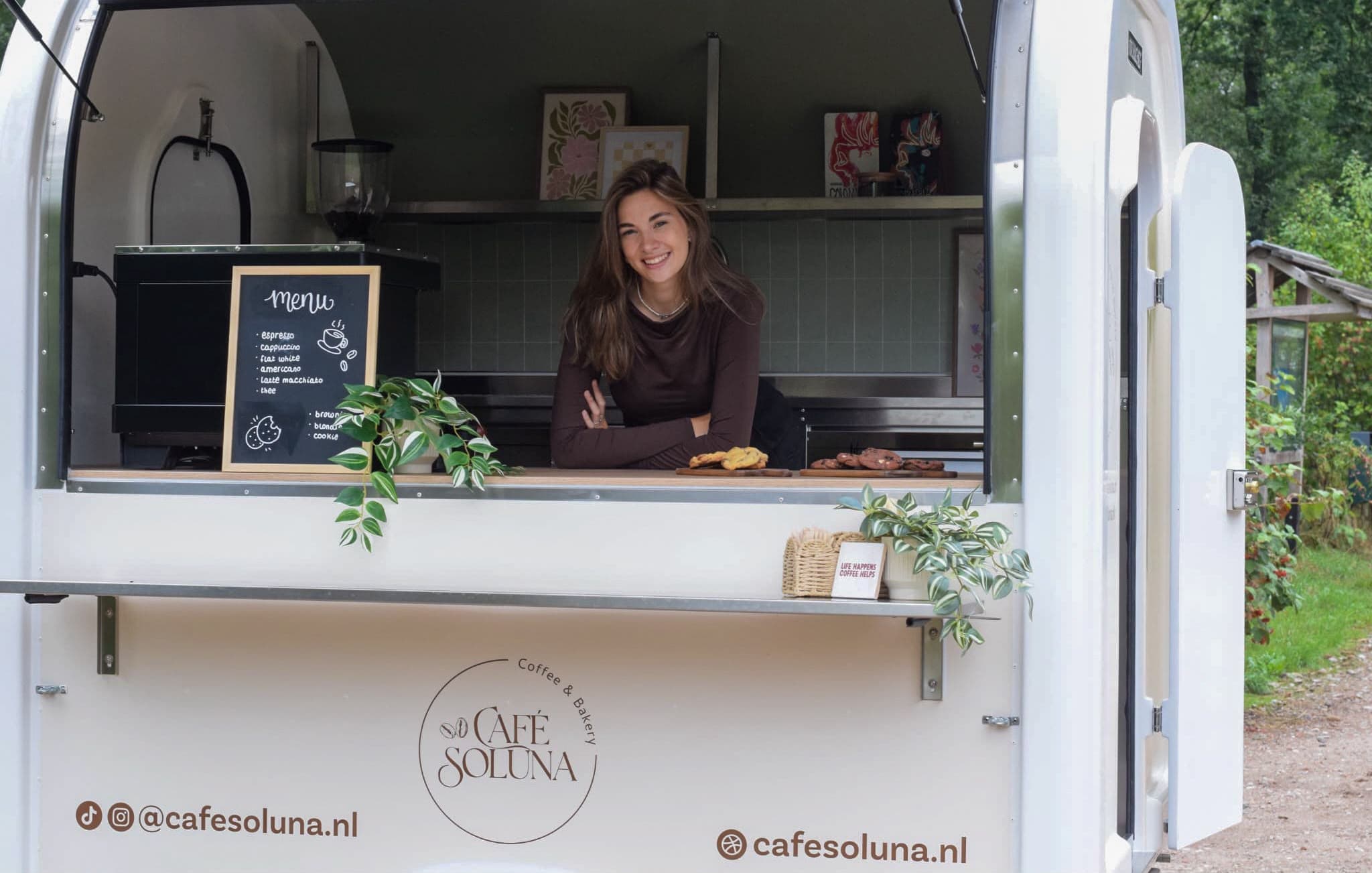 Melissa achter de toonbank in de geopende foodtruck van Café Soluna, met het menubord, vers gebak en het logo op de zijkant